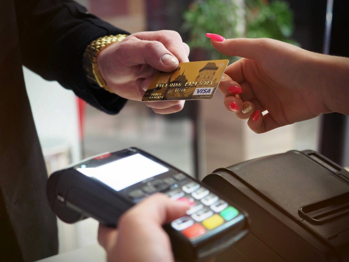 A black payment terminal is being held while a card is being handed over from the customer to the person holding the payment terminal