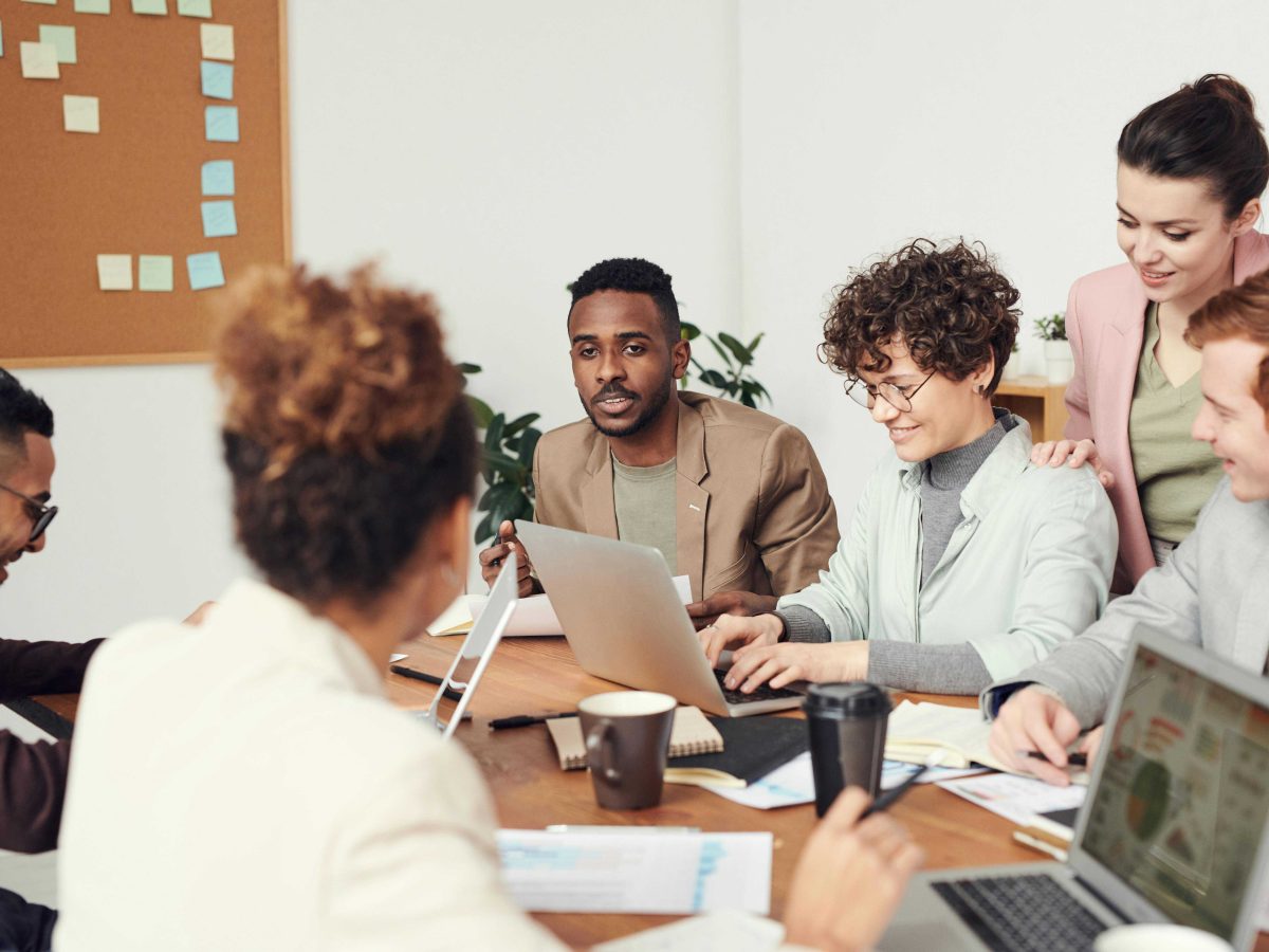 A team of six colleagues of various ethnicities working on a desk and communicating with each other