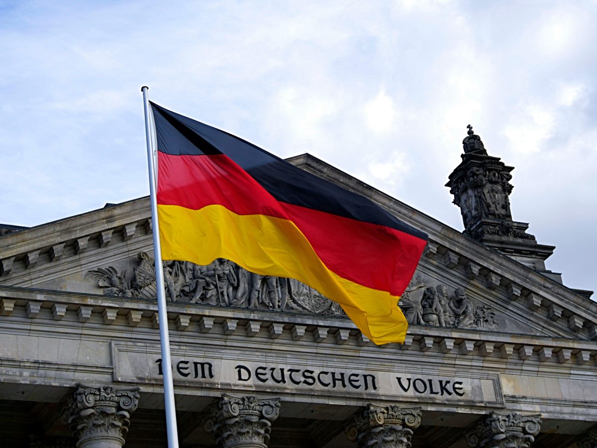 Photo of a German flag flying in the wind, with a historical building in the background.